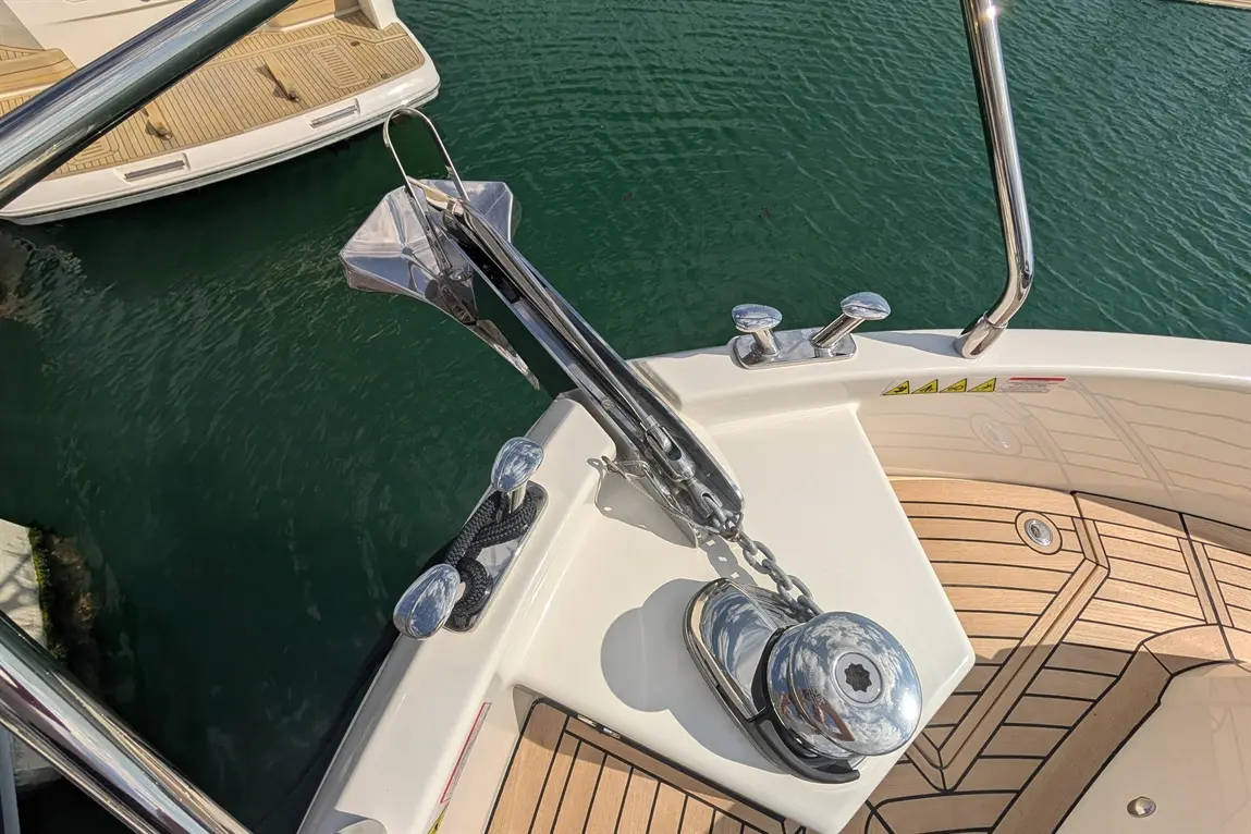 Boat bow with polished anchor, chain, and cleats on a wooden deck, above green water with another boat visible.